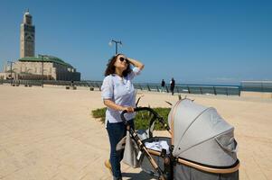 Young mother pushing baby pram in the Atlantic Ocean promenade, against the background of Mosque Hassan 2 in Casablanca photo