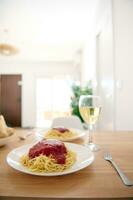 A plate with appetizing spaghetti and classic tomato sauce and a wine glass of white wine on the table at home interior photo