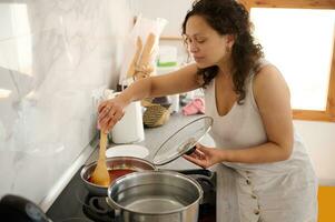 un ama de casa emocionante tomate passata en un cacerola, preparando salsa para italiano pasta en un minimalista hogar cocina foto