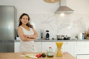 confidente encantador joven mujer con su brazos doblada, en pie por cocina, sonriente lindo mirando a cámara. foto