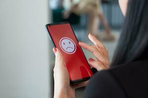 A woman holding a smartphone with a bad feeling emoticon on screen at a restaurant. concept of evaluating customer satisfaction on service levels and recommending that stores should improve. photo