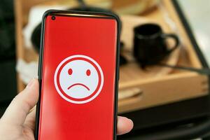 A woman holding a smartphone with a bad feeling emoticon on screen at a restaurant. concept of evaluating customer satisfaction on service levels and recommending that stores should improve. photo