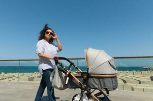 Beautiful woman enjoying a promenade along Atlantic seacoast in warm sunny day, pushing a stroller with her newborn baby photo