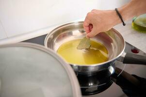 Close-up of a man's hand holding a bay leaf and seasoning the olive oil frying in a pan while cooking dinner at home photo