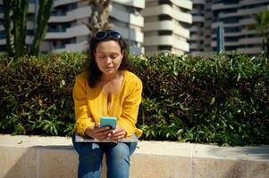 Charming mixed race woman using smart phone, checking new mobile app, browsing web sites, sitting outdoor photo