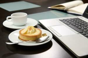 es hora a tomar un café descanso. un taza de recién elaborada Café exprés y dulce postre cerca un ordenador portátil. negocio concepto foto