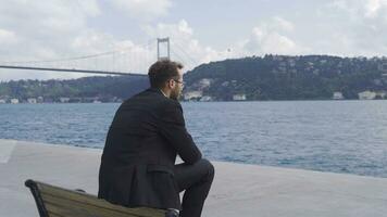 Thoughtful businessman sitting on bench by the sea in the city. video