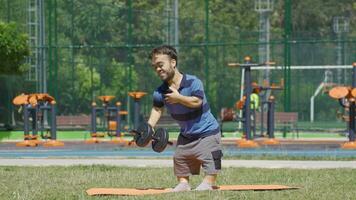 Dwarf young man trying to develop back muscles using dumbbells outdoors. video