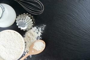 Baking ingredients flour, eggs, milk, bakeware on black slate background with copy space photo