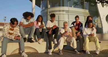 Big group of happy teenage friends each one looking at their phone in a bench in the street. video