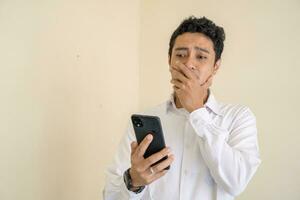 indonesio hombre vestir blanco ropa sorprendido cuando mirando el teléfono inteligente foto