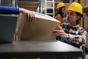 Storehouse order picker in wheelchair doing order fulfillment and taking cardboard box from coworker hands. Asian man with disability managing postal service operations in industrial warehouse photo