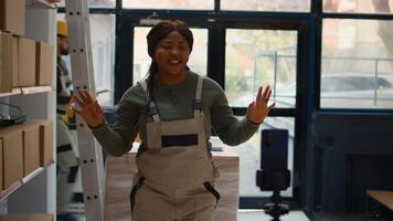 Warehouse chief supply chain officer using smartphone to make training video for interns. African american supervisor films herself in depository showing trainees how to seal cardboard boxes photo