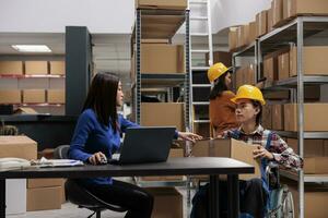 Warehouse manager coordinating logistics operators team from desk and checking parcel packing quality. Asian storehouse worker in wheelchair giving cardboard box to coworker at table photo