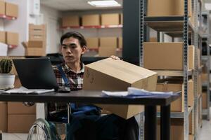 Asian wheelchair user managing parcel delivery in postal service warehouse. Storehouse shipment operator with disability holding cardboard box with customer order and planning shipping route on laptop photo