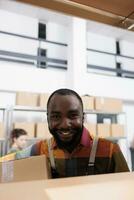 Smiling african american employee working at clients order, putting products in cardboard boxes, preparing products for shipping. Supervisor wearing industrial overall while carrying packages photo
