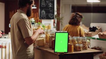 Client shops next to chroma key tablet with copy space used as commercial sign in zero waste supermarket. Promotional ad on green screen device in food store with products in nonpolluting package video