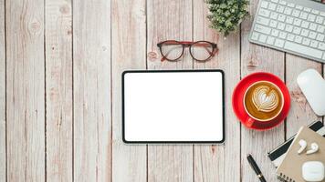 Wooden desk workplace with blank screen tablet, keyboard, mouse, eyeglass, notebook, pen and cup of coffee, Top view flat lay with copy space. photo