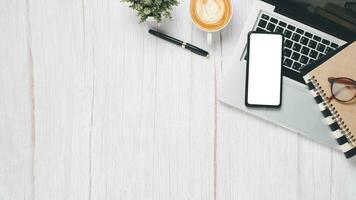 White wooden desk workplace with blank screen smart phone, laptop, pen, eyeglass, notebook and cup of coffee, Top view flat lay with copy space. photo