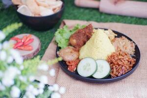 indonesian style yellow rice with chicken and side dishes in black plate on green grass background photo