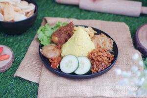indonesian style yellow rice with chicken and side dishes in black plate on green grass background photo