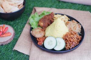 indonesian style yellow rice with chicken and side dishes in black plate on green grass background photo