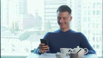 Cheerful handsome man smiling joyfully, using his smart phone, drinking tea video