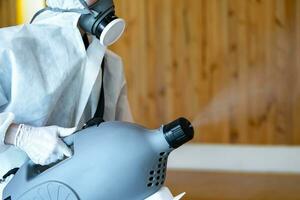 un técnico profesional con traje de prevención rocía una solución esterilizante con una máquina de pulverización eléctrica en el suelo de madera y fondo blanco con luz de estudio. foto