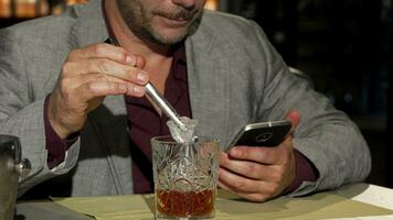 volwassen Mens gebruik makend van slim telefoon terwijl toevoegen ijs kubussen naar zijn whisky video