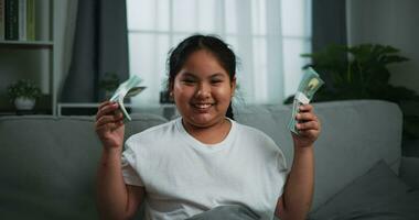 imágenes de alegre adolescente niña demostración dólar a cámara y sonriente en sofá en el vivo habitación a hogar. video