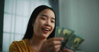 Aufnahmen von jung asiatisch Frau zeigen Dollar beim Kamera und lächelnd auf Sofa im das Leben Zimmer beim heim. video