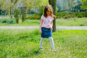 hermosa pequeño niña con desarrollando pelo camina en calle violando cuarentena de coronavirus. niña en rosado camiseta carreras a través de verde campo. niña obras de teatro activo juegos en naturaleza en verano o primavera. foto