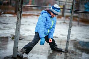 un niño en botas de nieve botas es en pie en un charco de Derretido nieve. hielo en el carreteras en el ciudad. no limpiado desde nieve y hielo territorio. mojado pies en invierno. foto