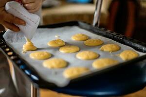 mano apretando fuera de confitería bolso masa para lactoso pasteles lactoso masa delicioso crema soplo pasteles en horneando bandeja. lactoso pasteles Profiteroles antes de colocación en horno foto