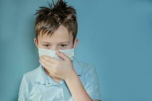 boy in medical protective mask teenager covers mouth with hand and coughs. on blue background. child with flu, influenza or cold protected from viruses, pollution, among patients with coronavirus. photo