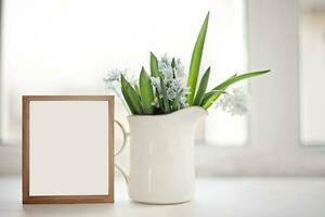 White ceramic jug with Fresh blue spring flowers Scilla siberica on windowsill on background of window. wooden empty frame with white background. Copy space photo