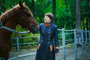 A woman in a long blue coat strokes the horse's face against the background of a green forest. A woman stands at an iron fence enclosing a pen for horses. photo