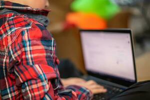 boy in plaid shirt does lessons on laptop. child learns remotely during quarantine of coronavirus. Online learning from home. photo