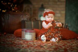 pequeño niña en rojo vestir en contra antecedentes de Navidad árbol sostiene Navidad guirnalda en su manos. bebé 6 6 mes antiguo celebra Navidad. foto