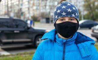 boy in face mask on street near road with cars. Child walks during quarantine and lockdown. child protected from viruses, pollution in bad epidemic situation, among patients with coronavirus photo