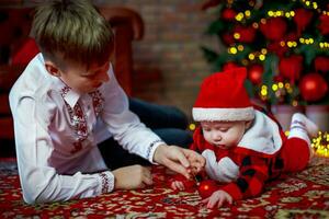 hermano con pequeño hermana a el nuevo año árbol. familia celebra Navidad. 6 6 meses antiguo pequeño chico aprende a gatear foto