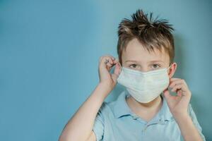 boy removes protective medical mask from face on blue background. child with flu, hantavirus or cold protected from viruses, pollution in bad epidemic situation, among patients with coronavirus photo