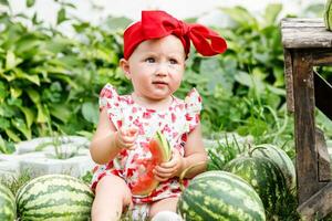 slice of ripe watermelon in the hands of a little toddler girl sitting near a pile of watermelons at a fair on a sunny summer day. Fresh fruit snacks. photo