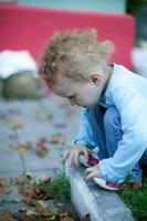 pequeño bonito niña con corto Rizado pelo se sentó allanamiento y mirando abajo. niño en pantalones y un azul camisa jugando al aire libre foto