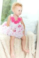 Sad little girl sitting on a window sill. A child in a smart dress is sitting on a soft blanket near the pillow. photo