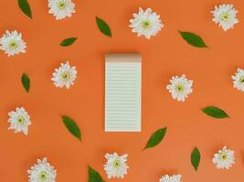 Directly above view of blank notebook with white flowers and green leafs on orange background. photo