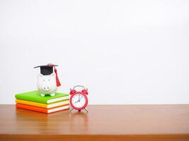 Piggy bank with graduation hat on hardcover book and red alarm. Copy space for text, Back to school, Education concept photo