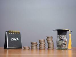 Study goals, 2024 Desk calendar, Glass bottle with graduation hat and stack of coins. The concept of saving money for education, student loan, scholarship, tuition fees in New Year 2024 photo