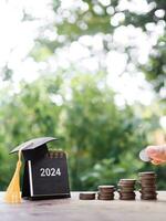 Close up hand putting coin in stack of coins, 2024 Desk calendar with graduation hat. The concept of saving money for education, student loan, scholarship, tuition fees in New Year 2024 photo