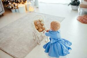 Baby girl wearing cute dress and headband, carries stroller in festively decorated room with garland of lights. photo